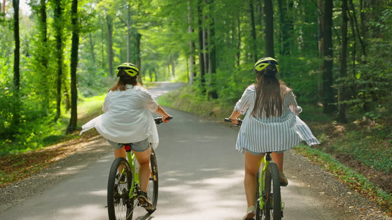 Two people wearing helmets riding green bicycles on a paved path through a sunlit forest