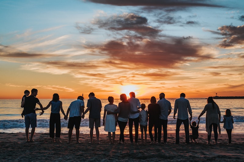 Large family standing silhouetted on a sandy beach holding hands while facing an orange sunset over the ocean